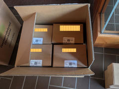 An open box placed inside a building, containing smaller labeled cardboard boxes neatly arranged in rows. A door with glass panels is visible in the background.