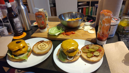Auf einem vollen Tisch werden zwei Double-Cheeseburger auf Tellern angerichtet, mit Brötchen, die mit Soße bestrichen und mit Salat, Zwiebelringen, Gurkenscheiben und geschmolzenem Käse belegt sind. Im Hintergrund sind ein Schneidebrett mit geschnittenen Zwiebeln und Kirschtomaten, eine Schüssel mit Pommes frites sowie verschiedene Gewürze und Flaschen zu sehen.