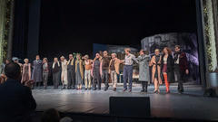 Darsteller in verschiedenen Bühnenkostümen stehen während des Vorhangs Arm in Arm auf einer Theaterbühne, vor einem stilisierten Bühnenbild aus Zylindern, in denen sie spielten, und mit Silhouetten des Publikums im Vordergrund.
