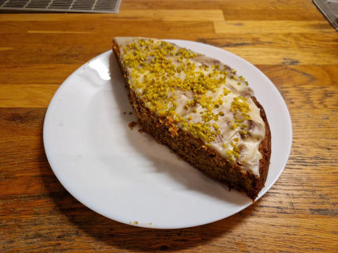A slice of cake on a white plate, topped with white icing and chopped pistachios, sitting on a wooden countertop.