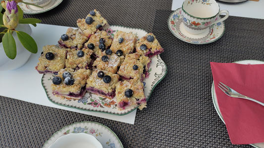Eine mit Blumen verzierte Servierplatte, gefüllt mit Blaubeer-Streuselkuchen-Quadraten, bestäubt mit Puderzucker, auf einem Tisch mit gemusterten Tischsets, einer Teetasse und Untertasse sowie einer gefalteten roten Serviette mit einer Gabel.