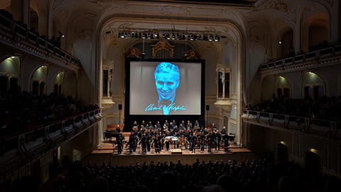 Wide view of an ornate concert hall with a seated audience and an orchestra on stage beneath a large screen showing a blue-tinted portrait and a signature of Charlie Chaplin.