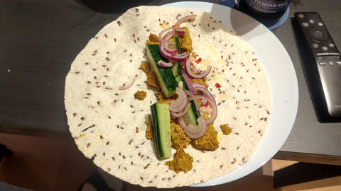 Ansicht auf einen offenen Tortilla-Wrap mit Körnern auf einem Teller, gefüllt mit Gurkensticks, geschnittenen roten Zwiebeln und zerbröckelter Gewürzfüllung, mit einer Fernbedienung neben dem Teller.