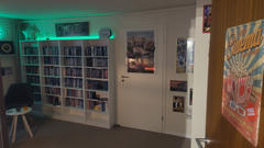 Ein gemütlicher Heimkino-Raum mit einem großen Regal voller DVDs/Blu-rays, beleuchtet von grüner LED-Beleuchtung, Filmplakaten an den Wänden und der Tür sowie einem farbenfrohen „Cinema“-Poster mit Popcorn-Motiv an einer nahegelegenen Tür.