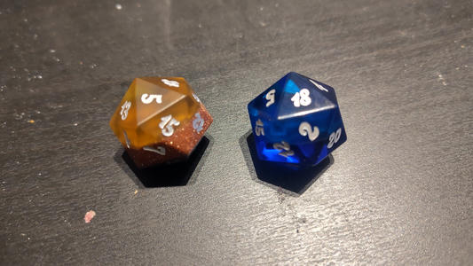 Two 20-sided dice on a dark tabletop—one translucent amber with glitter and white numbers, and one translucent blue with white numbers.
