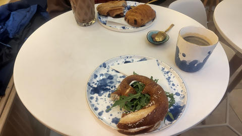 Kleines rundes Café-Tischset mit Gebäck und Getränken, darunter ein Brezel-Sandwich auf einem gemusterten Teller, ein Schoko-Croissant und Gebäck im Hintergrund sowie eine Tasse Kaffee + heiße Schokolade.