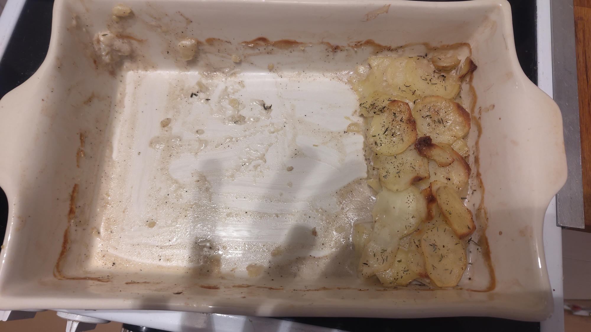 A mostly empty ceramic baking dish with browned edges and a small portion of sliced, baked potatoes remaining in one corner.