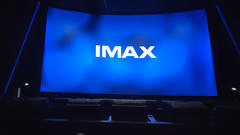 Inside a dark movie theater, a large screen displays the word “IMAX” in white on a bright blue background, with seats visible in the foreground.