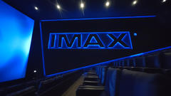 Inside an IMAX theater with rows of dark seats and blue accent lighting, the large screen shows the “IMAX” logo.