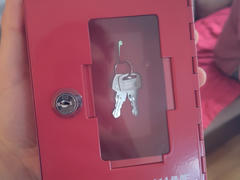 Eine rote, abschließbare Aufbewahrungsbox mit einem transparenten Fenster, hinter dem zwei silberne Schlüssel an einem Ring zu sehen sind. Die Box wird von jemandem in der Hand gehalten, und hinter ihr ist ein Teil des umgebenden Raums zu sehen.