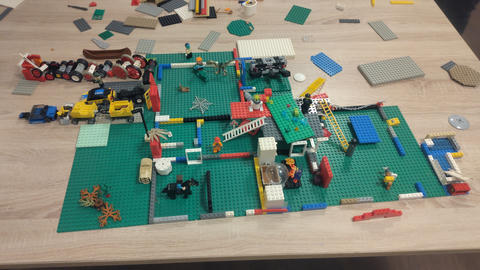 A tabletop LEGO scene built on large green baseplates, with scattered loose bricks and pieces around it, plus small vehicles, minifigures, and structures.