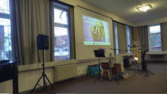 Kleiner Veranstaltungsraum mit einer Projektionswand, auf der eine deutsche Präsentationsfolie („Bis auf die Knochen“) gezeigt wird, davor stehen Mikrofone, Notenständer, Lautsprecher und Percussion-Instrumente.