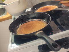 Zwei Bratpfannen auf einem Herd, in denen goldbraune Pfannkuchen gebacken werden. Eine weiße Rührschüssel steht auf der Arbeitsplatte daneben, im Hintergrund ist eine etwas unordentliche Küche zu sehen.