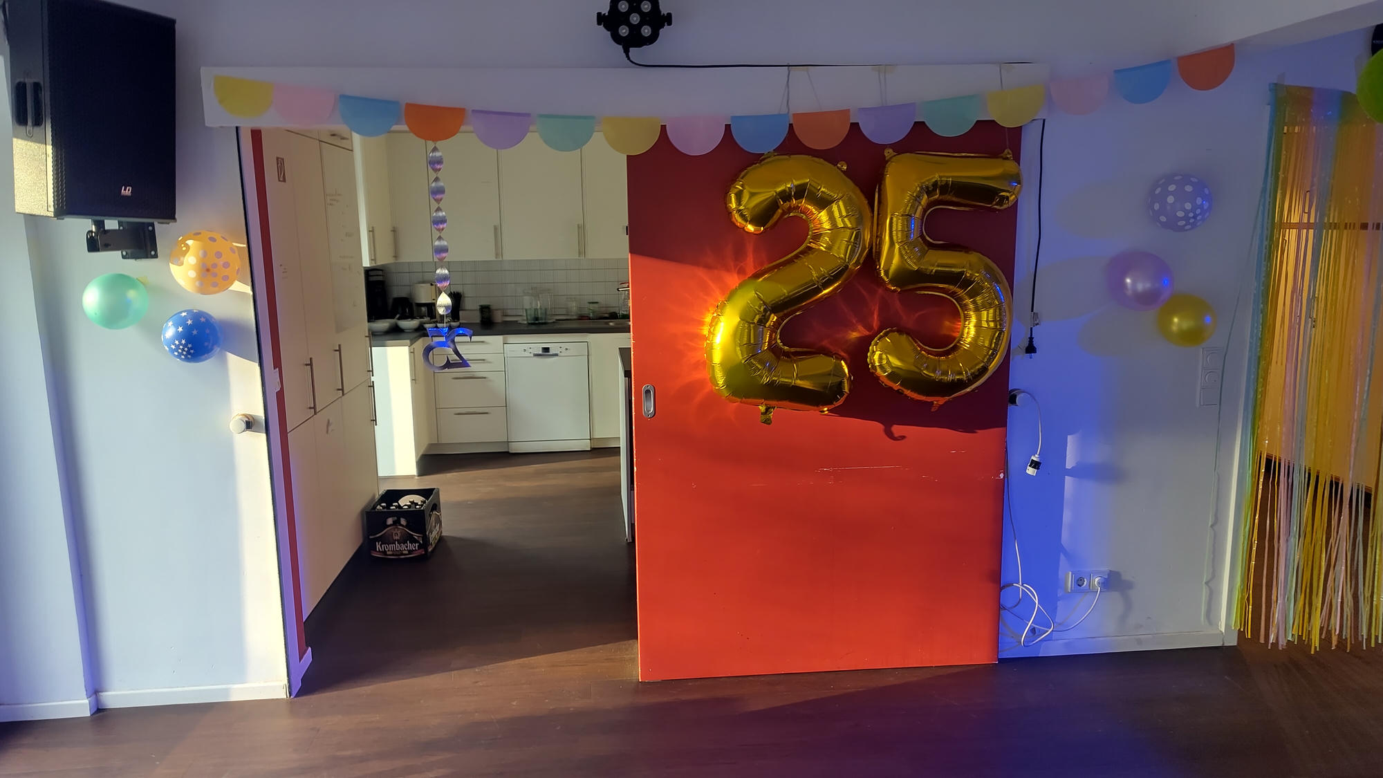A decorated party room with colorful bunting and balloons; large gold “25” number balloons are attached to a red sliding door in front of a kitchen area.