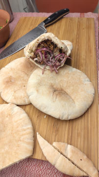 Pita breads on a wooden cutting board, with one pita cut open and stuffed with seasoned filling and sliced red onions; a chef’s knife lies behind it.