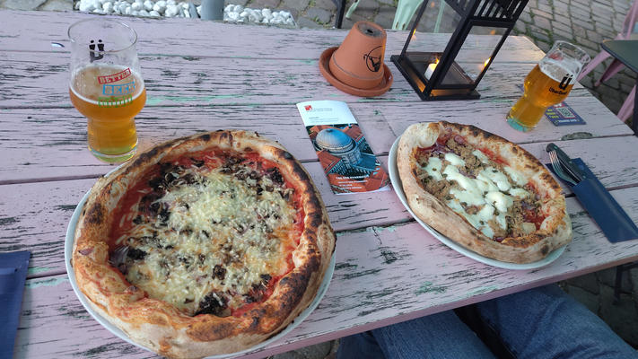 Two unique pizzas and two glasses of beer sit on a weathered pink outdoor table. A small lantern, an upside-down terracotta pot, and a brochure are placed between the pizzas, with cutlery resting to the side.