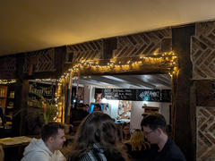 A cozy pub interior with warm lighting and brick walls + thick wooden structures. A very old building. String lights frame a wooden doorway leading to a brighter room with a chalkboard menu and a bulletin board. Patrons are seated at tables, enjoying their time.