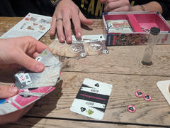 A tabletop scene where people are playing the card game Staying Alive. Hands are visible holding cards, and there are game cards, tokens, a sand timer, and a partially consumed pint of beer on the rustic wooden table.