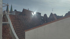 Blick vom Dach auf rotgedeckte Gebäude im Sonnenlicht, mit Kirchtürmen in der Ferne und einer hellen Sonnenreflexion auf einem Dachfenster.