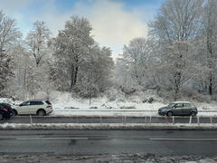 A view of a snowy roadside landscape with cars along the street and snow-dusted trees in the background. The sky is partly cloudy with patches of blue visible.