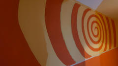 Sieht aus wie eine geschwungene Flurwand, gestrichen in kräftigem Orange und mit weißen spiralförmigen Streifen (ein Schatten mit gelben Effekten ist zu sehen), die eine optische Täuschung einer Kurve erzeugen, da es sich um eine flache Wand handelt.
