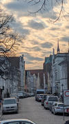 Kopfsteinpflasterstraße, gesäumt von geparkten Autos und schmalen historischen Gebäuden, die zu einer großen Kirche mit einem Turm unter einem bewölkten Abendhimmel führt.