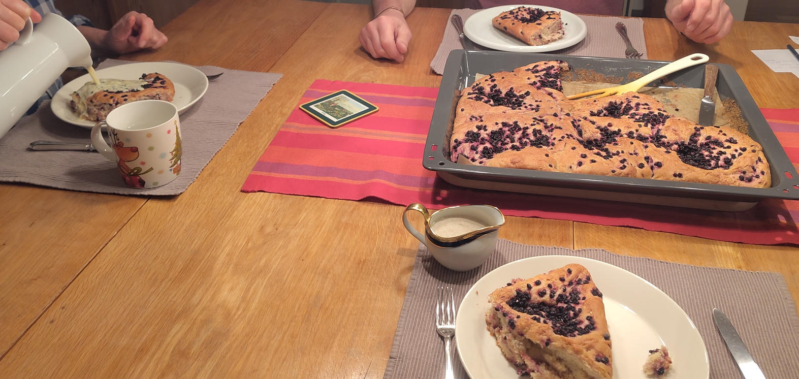 Ein Bild des schwedischen Kuchens in der Mitte eines Esstisches, mit drei Stücken auf Tellern. Auf einem davon gibt eine Person (nur ihre Hände sind zu sehen) die Vanillesauce darauf