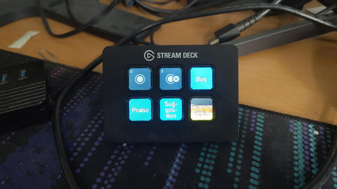 Close-up photo of an Elgato Stream Deck Mini on a desk, with six illuminated buttons including “Bug,” “Praise,” “Suggestion,” and “German Nitpick,” surrounded by cables.