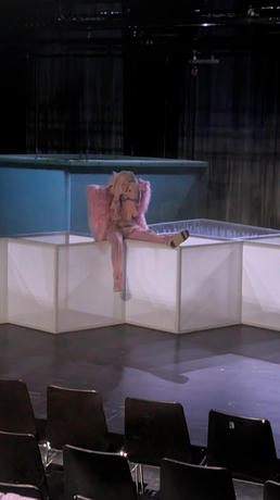 A theater stage featuring a person dressed in an pig stuffed animal. They are sitting with one leg extended on a white geometric structure, and empty black chairs can be seen in the foreground.