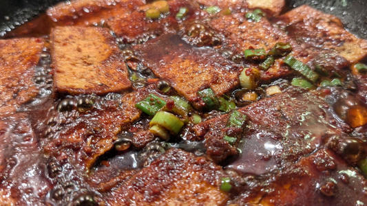 Close-up of food simmering in a dark, glossy sauce, with browned rectangular tofu pieces and chopped green onions visible on the surface.