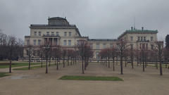 A large, pale stone palace building viewed from a formal garden with rows of leafless trees and gravel paths under an overcast sky.