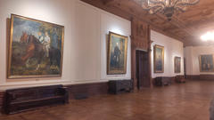 An elegant gallery room with wood-paneled ceiling and chandeliers, showing several framed portrait paintings on white walls above wooden benches.