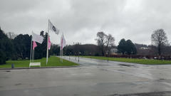 A wet road curves through a landscaped park with grass lawns and tall flagpoles flying white and pink flags under a gray, overcast sky.