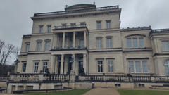 Front view of a large neoclassical stone mansion with tall columns, many windows, and a balustrade, photographed on a cloudy day.